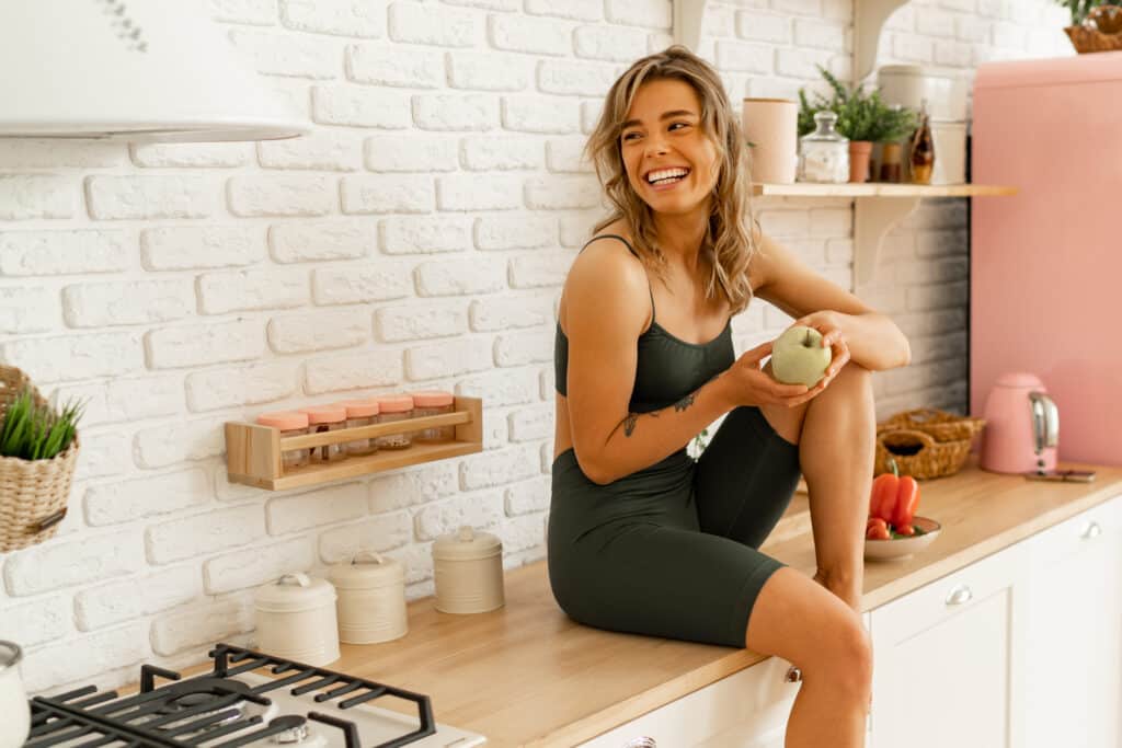Laughing blond woman holding apple and posing on light kitchen. Healthy food concept.