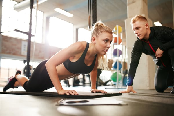 Blonde woman in gym on hands and knees doing a pushup with a trainer next to her